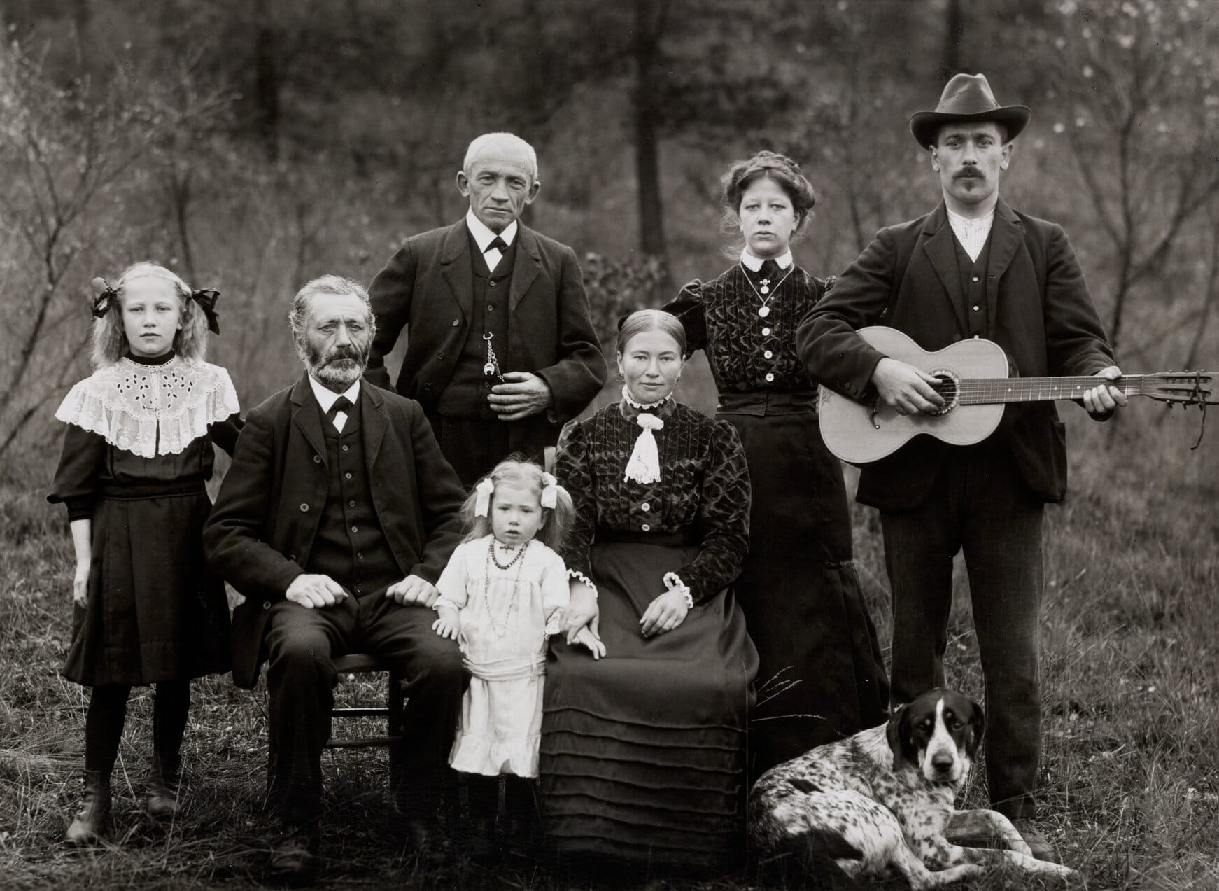 A black-and-white photograph of a group of people posing for the camera outdoors. Two adults are seated toward the center, alongside two standing children. Three adults stand behind them. The man at far right holds a guitar, with a dog laying at his feet. The men wear three-piece suits and the women long skirts with textured or patterned tops.
